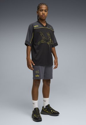 Ung person står iført sort og gråt Pokemon-tema Puma sportstøj med Pikachu-designs og sorte og gule sneakers.