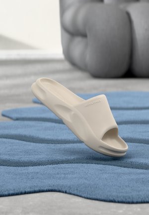 Beige skumsandalett med öppen tå, böjda kanter och en texturerad fotbädd, visad på en blå texturerad matta.