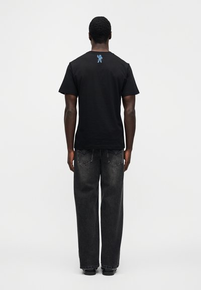 Μαύρο βαμβακερό μπλουζάκι με κοντά μανίκια και μπλε κεντητό σχέδιο στο πάνω μέρος της πλάτης, συνδυασμένο με άνετο σκούρο γκρι τζιν παντελόνι.