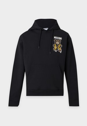 Mladý muž stojaci, oblečený v čiernej mikine s kapucňou a nášivkou medvedíka, čiernych nohaviciach so zlatým zipsom, čiernych topánkach a bielom tričku.