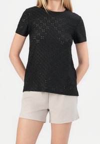 Camisa negra de manga corta hecha de una tela texturizada con patrones calados. Combinada con pantalones cortos beige, mostrando un ajuste relajado y un diseño casual.