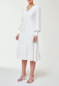 Biała sukienka z długim rękawem i dekoltem w kształcie litery V, z haftowanymi akcentami i falbanistym dołem. Gładki materiał z luźnym krojem, w połączeniu z białymi sandałami.
