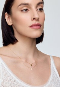 Złote, warstwowe naszyjniki z pendetem w kropelce i owalnym, noszone na nagiej szyi, z lekką, teksturowaną białą odzieżą.