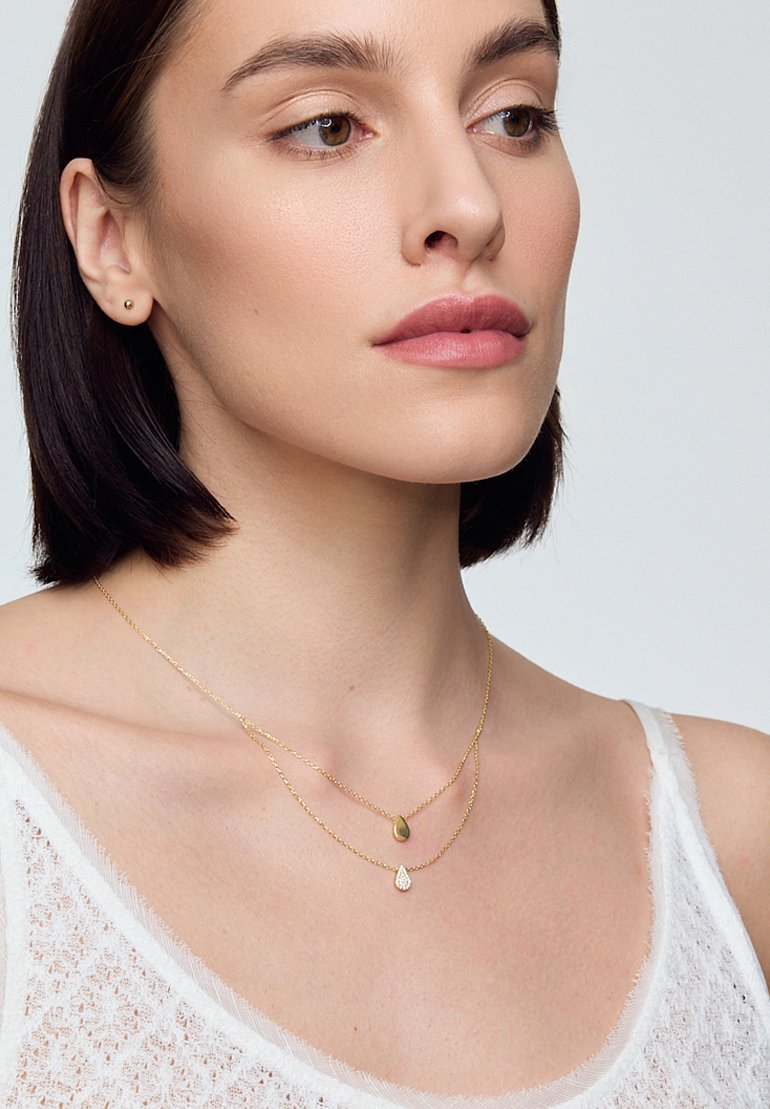 Złote, warstwowe naszyjniki z pendetem w kropelce i owalnym, noszone na nagiej szyi, z lekką, teksturowaną białą odzieżą.