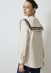 Crème blouse met een contrasterende donkere kraag. De kraag heeft twee strepen. Lange mouwen met knoopmanchetten. Gladde textuur, casual fit.