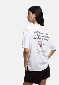 Witte katoenen t-shirt met korte mouwen, met een teddybeer graphic en tekst op de achterkant. De stof lijkt zacht met een relaxte pasvorm.