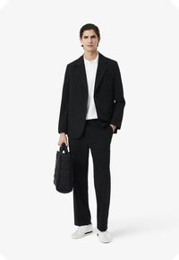 Blazer nero su misura sopra una polo bianca, abbinato a pantaloni neri. Porta una borsa trapuntata nera, completando il look con sneakers bianche.