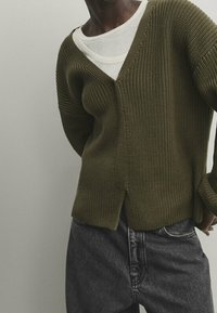 Persona che indossa un cardigan lavorato a maglia verde oliva sopra una maglietta bianca a girocollo e jeans alti di colore grigio scuro, in piedi con le mani dietro la schiena.