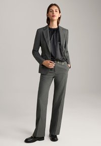 Grauer maßgeschneiderter Anzug mit strukturierter Jacke und weitem Hosenbein. Schwarzes Satinoberteil mit Rüschenausschnitt. Schwarze Schuhe mit metallischen Akzenten.
