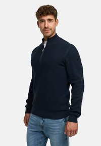 Marineblauer Pullover mit halbem Reißverschluss, geripptem Textur und schmalen Ärmeln. Kombiniert mit hellblauen Jeans, die ein dezentes Logodesign aufweisen.