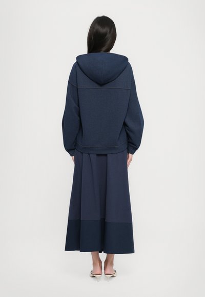 Sudadera marinera con una gran capucha y dobladillo acanalado, combinada con una larga falda marinera fluida que tiene un panel inferior más oscuro. Textura suave.