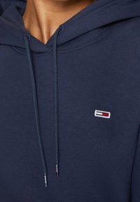 Gros plan d'une personne portant un sweat à capuche bleu marine avec des cordons et un petit logo rectangulaire rouge, blanc et noir sur la poitrine.