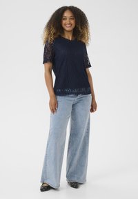 Top in pizzo blu navy con maniche corte e orlo scallop, abbinato a jeans a gamba larga blu chiaro e mule nere con tacco e borchie.