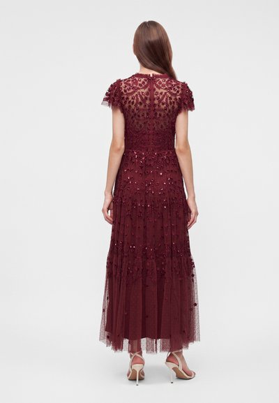 Femme aux longs cheveux bruns portant une robe bordeaux en dentelle et paillettes avec manches courtes et dos transparent, associée à des sandales blanches à talons hauts.
