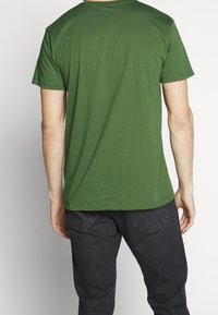 Man med en enfärgad grön t-shirt med kort ärm och svarta jeans, sedd bakifrån mot en plain ljus bakgrund.