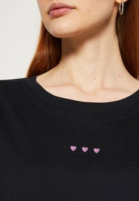 Gros plan d'une personne portant une chemise noire avec trois petits symboles de cœur roses centrés sur la poitrine.