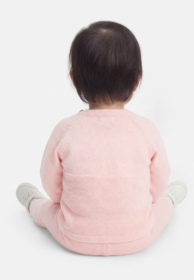 Νήπιο καθισμένο σε σταυροπόδι, γυρισμένο προς τα πίσω, φοράει απαλό ροζ πλεκτό σύνολο και λευκά παπούτσια, πάνω σε απλό λευκό φόντο.