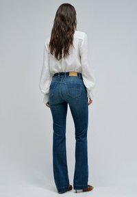 Mujer con pelo largo y ondulado que lleva una blusa blanca de manga larga, jeans de cintura alta azules y zapatos de tacón marrones, de espaldas.