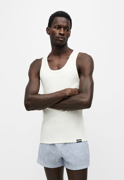 Homme les bras croisés portant un débardeur blanc et un short rayé bleu clair, debout devant un fond uni clair.