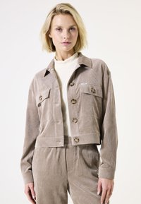 Bruine corduroy jas met knoopsluiting, twee voorzakken en een cropped ontwerp. Draagt over een gestructureerde lichte top. Gladde textuur.