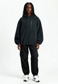 Giacca in pile nero con chiusura a zip frontale, vestibilità oversize e tasche laterali. Abbinata a pantaloni cargo neri e sneakers bianche.