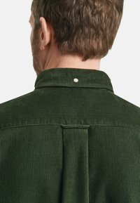 Camisa de pana verde con cuello abotonado, estriado verticalmente y un pliegue en la parte trasera para añadir textura y estructura.