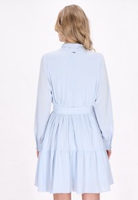 Hellblaues Kleid mit einem Rüschensaum, langen Ärmeln und einem Gürtel. Glattes Material mit einem Kragen und dezentem Knopfdetail auf der Rückseite.