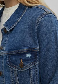 Modrá džínová bunda s knoflíkovým zapínáním, špičatým límcem a náprsní kapsou s výrazným obnošeným detailem.