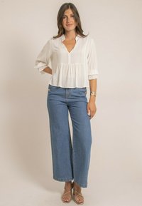 Blouse blanche texturée avec un décolleté en V et des manches courtes à volants, associée à un jean en denim bleu à taille haute et à jambes larges, ainsi qu'à des sandales à talons.