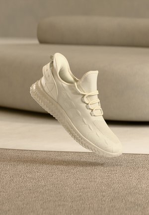 Scarpa da ginnastica atletica beige chiaro con tomaia in maglia testurizzata, suola ammortizzata e design senza lacci, posizionata sopra un pavimento moquettato davanti a un divano beige.