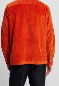 Camicia in corduroy arancione con righe verticali, polsini abbottonati e orlo dritto. Tessuto testurizzato con una finitura liscia e cuciture visibili.