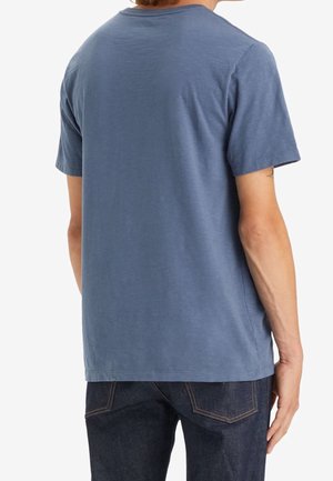 Personne portant un t-shirt bleu uni à manches courtes et un jean bleu foncé, vue de dos sur un fond blanc.