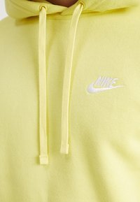 Žlutá mikina s kapucí vyrobená z měkké tkaniny, s bílým logem Nike a šňůrkami na kapuci. Hladký povrch bez vzorů.