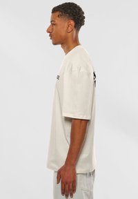 Beige Baumwoll-T-Shirt mit kurzen Ärmeln, lockerer Passform und schwarzem Text auf der Brust. Seitenansicht zeigt einen geraden Saum und leichte Textur.