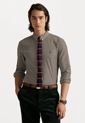Camicia a righe con logo verde, colletto stretto; abbinata a una cravatta a motivo, cintura marrone e pantaloni in velluto a coste verde scuro. Orologio al polso.