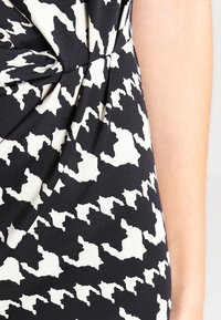 Czarna i biała tkanina w geometryczny wzór z detalem w formie skrętu; tekstura wydaje się gładka z delikatnym połyskiem; abstrakcyjne kształty tworzą dynamiczny wizual.