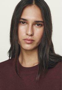 Maroon sweatshirt with a crew neckline, soft fabric, and straight cut. Model has long dark hair and neutral makeup, facing forward.