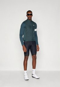 Fietsjack in donker teal met een ritssluiting aan de voorkant, lange mouwen en een witte accentstreep. Gecombineerd met zwarte shorts en witte sportschoenen.