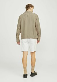Camisa de manga corta color oliva con dobladillo redondeado, combinada con pantalones cortos blancos. El modelo lleva zapatos negros y calcetines beige. Diseño minimalista.