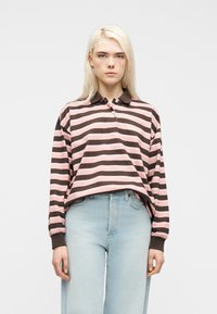 Νέα γυναίκα με μακριά ξανθά μαλλιά που φοράει ροζ και καφέ ριγέ μπλούζα πόλο με μακριά μανίκια και ανοιχτό μπλε ψηλόμεσα τζιν.