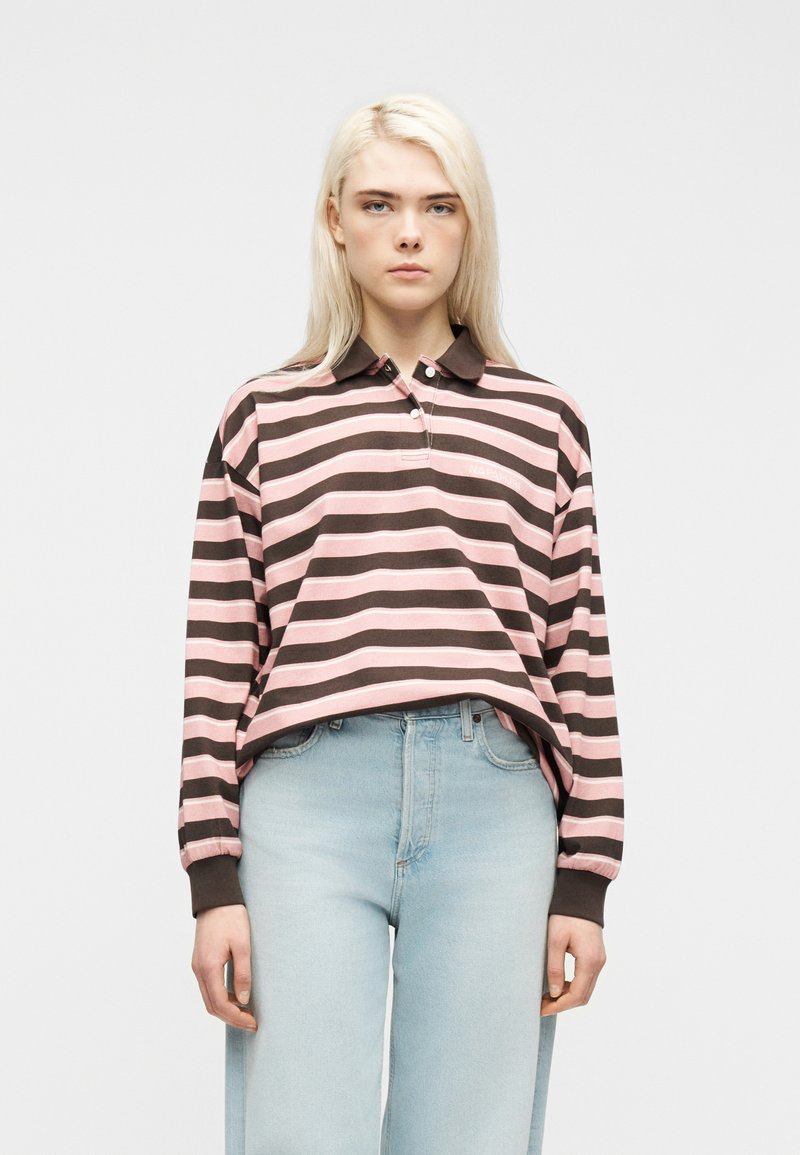 Νέα γυναίκα με μακριά ξανθά μαλλιά που φοράει ροζ και καφέ ριγέ μπλούζα πόλο με μακριά μανίκια και ανοιχτό μπλε ψηλόμεσα τζιν.