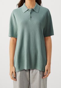 Polo à manches courtes vert clair, en tissu maille texturé, avec un col classique et trois boutons à l'avant. Associé à un pantalon gris clair.