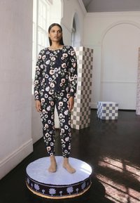 Femme se tenant sur une plateforme ronde peinte, portant un ensemble coordonné haut et pantalon à fleurs sombres avec des chaussettes beiges, dans une pièce lumineuse et minimaliste.
