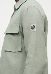 Ανοιχτό γκρι μακρυμάνικο πουκάμισο με μεγάλο τσεπάκι στο στήθος και μπλε τσόχα με το λογότυπο της Belstaff στο πάνω μέρος του μπράτσου, φορεμένο από ένα άτομο που φαίνεται από το πλάι.