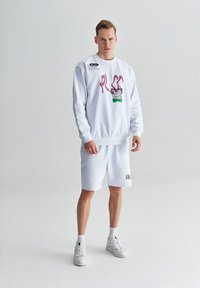 Biała bluza z różowym i zielonym graficznym wzorem, zestawiona z białymi szortami. Posiada ściągacze na mankietach i u dołu. Noszona z białymi sneakerami.