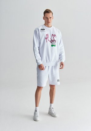 Biała bluza z różowym i zielonym graficznym wzorem, zestawiona z białymi szortami. Posiada ściągacze na mankietach i u dołu. Noszona z białymi sneakerami.