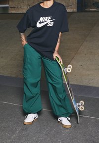 Osoba oblečená v černém tričku Nike SB, zelených cargo kalhotách a bílých sneakers držící skateboard uvnitř na šedé podlaze.