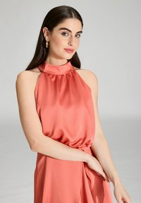 Donna con lunghi capelli scuri che indossa orecchini a cerchio dorati e un vestito di raso corallo senza maniche con scollatura alta, in posa contro uno sfondo neutro.