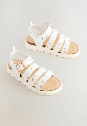 Hvide blomstrede sandaler med tre brede stropper, ankelspænde og beige polstret fodseng på en tekstureret hvid sål.