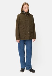 Olijfgroene, gevoerde jas met een hoge kraag en capuchon. Bevat twee voorkruiken en drukknoopsluitingen. In combinatie met blauwe denim en zwarte schoenen.